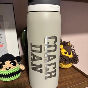 May include: A gray stainless steel water bottle with a black lid and a silver inscription that reads "COACH DAN".