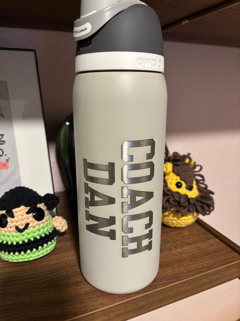 May include: A gray stainless steel water bottle with a black lid and a silver inscription that reads "COACH DAN".