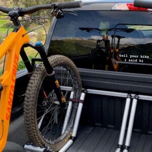 Peut inclure: Un v&eacute;lo de montagne orange vif avec un cadre noir et une fourche &agrave; suspension est s&eacute;curis&eacute; &agrave; l'arri&egrave;re d'une camionnette noire &agrave; l'aide d'un porte-v&eacute;los. La vitre arri&egrave;re du camion a un autocollant qui dit "dis &agrave; ton v&eacute;lo que je l'ai salu&eacute;".