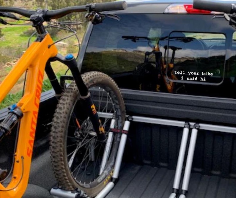 Peut inclure: Un v&eacute;lo de montagne orange vif avec un cadre noir et une fourche &agrave; suspension est s&eacute;curis&eacute; &agrave; l'arri&egrave;re d'une camionnette noire &agrave; l'aide d'un porte-v&eacute;los. La vitre arri&egrave;re du camion a un autocollant qui dit "dis &agrave; ton v&eacute;lo que je l'ai salu&eacute;".
