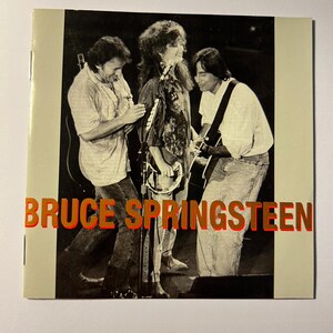 Puede incluir: Foto en blanco y negro de Bruce Springsteen actuando en el escenario con otros dos músicos. La foto está sobre un fondo crema con el texto "BRUCE SPRINGSTEEN" en letras naranjas.