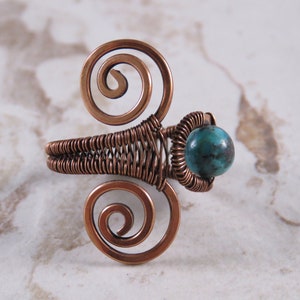 May include: A copper wire ring with a turquoise stone. The ring is made of two spirals on either side of the stone, and the band is woven with copper wire.