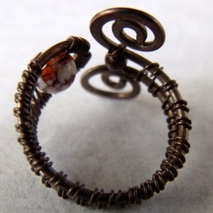 Op de afbeelding: Een ring van koperdraad met een witte en oranje steen. De ring is gemaakt van draad dat is gedraaid en om zichzelf is gewikkeld om een uniek ontwerp te cre&euml;ren.