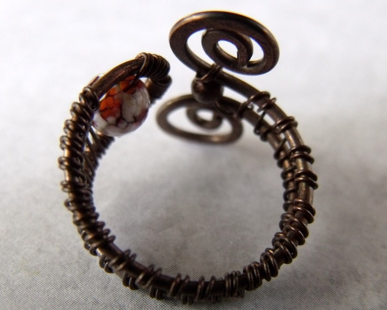 Op de afbeelding: Een ring van koperdraad met een witte en oranje steen. De ring is gemaakt van draad dat is gedraaid en om zichzelf is gewikkeld om een uniek ontwerp te cre&euml;ren.