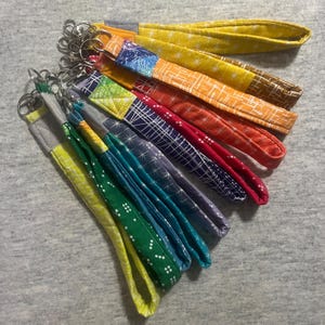 May include: A collection of colorful wrist keychains. Each keychain features a fabric wrist strap in various patterns and colors, including yellow, orange, red, blue, and green. The straps are attached to silver-toned key rings.