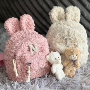 May include: Two fluffy bunny ear backpacks, one pink with an "N" and one cream with a "T". Two small teddy bear keychains and a white teddy bear are in front of the backpacks. The backpacks have zippered compartments.