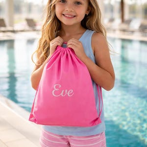 以下が含まれることがあります： 白い筆記体で「Eve」と書かれた明るいピンク色の巾着袋。明るい青色のタンクトップとピンクのストライプのショートパンツを着た人が持っています。背景にはプールとヤシの木があります。