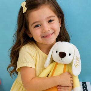 May include: A plush bunny toy with long ears, a white body, and a yellow outfit. The bunny has a brown nose and the name "Lily" embroidered on one ear. The toy is being held by a child wearing a yellow dress.