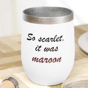 Puede incluir: Vaso de vino sin pie blanco con borde plateado y el texto "So scarlet, it was maroon" en letras negras y granates. Un sacacorchos y una tapa son visibles en primer plano.