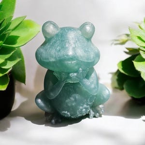 May include: A teal green frog figurine with a shiny, glittery finish. The frog is sitting with its hands clasped in front of it, as if in meditation.