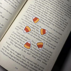 May include: Candy corn stickers scattered on an open book page. The stickers are white, orange, and yellow, with a classic candy corn design. The book's pages are yellowed, and the text is visible.