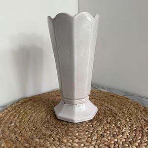 May include: A white ceramic vase with a scalloped rim and a geometric, faceted body. The vase has a pedestal base and stands on a round, woven mat. The vase is a neutral color and would complement many decor styles.