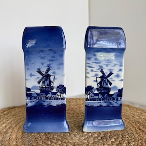 May include: Two blue and white ceramic vases with a Dutch windmill design. The rectangular vases feature a dark blue top and base, with a detailed landscape scene of a windmill, houses, and trees in the center. The vases are displayed on a woven mat.