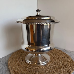May include: A silver-colored ice bucket with a lid. The bucket has a wide, fluted rim with a rope-like design. The lid is topped with a black knob. The bucket sits on a round base. The bucket is on a woven mat.