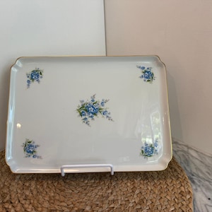 May include: Rectangular white serving tray with a gold trim and blue floral design. The tray features clusters of blue flowers in each corner and a larger arrangement in the center. The tray is displayed on a white stand.