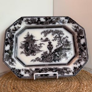May include: An octagonal, antique serving platter with a black and white Asian-inspired landscape design. The plate features pagodas, trees, and floral accents. The rim has a decorative border. The platter is displayed on a white stand.