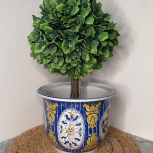 May include: A spherical artificial green topiary plant with a brown stem, potted in a round ceramic planter. The planter features a vibrant blue background with white vertical stripes and decorative oval panels containing yellow and blue floral motifs.