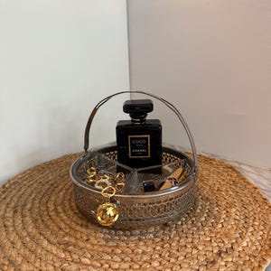 May include: A silver-toned metal basket with a handle, containing a black perfume bottle, gold jewelry, and a lipstick. The basket sits on a round, woven mat. The perfume bottle has the text "Coco Chanel".