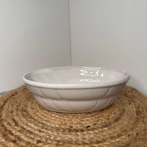 May include: A white ceramic serving bowl with a ribbed design around the base. The bowl sits on a round, woven, natural-colored placemat. The background is a plain white wall.
