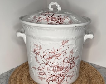 May include: A white ceramic lidded jar featuring a maroon floral transferware pattern. The cylindrical vessel has two side handles and a matching lid with a central knob. The surface displays embossed detailing around the rim and base.