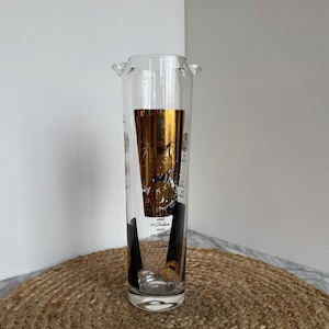 May include: A clear glass cocktail shaker with gold and black accents. The shaker features a gold foil design and black vertical stripes. The text on the shaker reads "1865 20 Dollars, 1829 2 Dollars, 1829 1 Dollar."