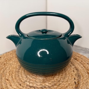May include: A dark teal ceramic teapot with a glossy finish, featuring a rounded body with horizontal ridges at the base, a matching lid with a small knob, and a wide arched handle that connects to the two symmetrical spouts on each side.