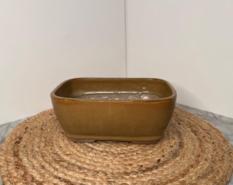 May include: A square, ceramic planter with rounded corners. The planter is a warm, earthy brown color with a glossy finish. It sits on a small, woven jute mat. The planter is designed for indoor or outdoor use.