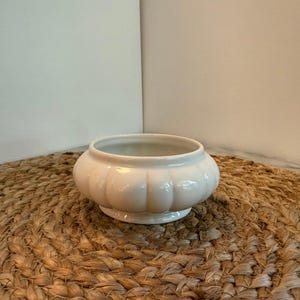 May include: A small, white ceramic bowl with a scalloped design around the exterior. The bowl sits on a woven, brown mat. The bowl has a rounded shape and a smooth, glossy finish. It is likely used for decorative purposes or as a small planter.