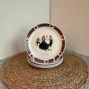 May include: A stack of white ceramic plates with a decorative border and a central image of people dancing in traditional attire. The plates are displayed on a woven mat.