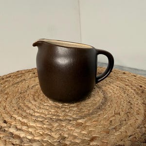 May include: A dark brown ceramic creamer pitcher with a smooth, matte finish. The pitcher features a rounded body, a curved handle on the right side, and a small, integrated pouring spout at the top rim, revealing a light beige interior.