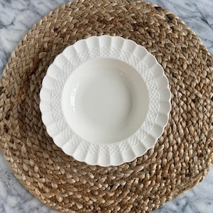 May include: A white ceramic bowl with a decorative rim sits on a round, woven placemat. The bowl has a scalloped edge and a textured band near the rim. The placemat is made of natural fibers and has a circular shape.
