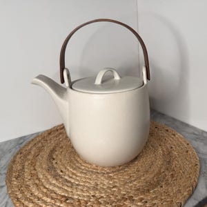 May include: A matte off-white ceramic teapot with a rounded body, a curved spout, and a matching lid with a small handle. The teapot features a dark brown, arched wooden handle attached to the sides, creating a contrast with the light ceramic.