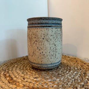 May include: A cylindrical ceramic vase with a speckled beige body and blue accents. The top and bottom feature horizontal blue bands, and the rim is a darker blue. The vase sits on a round, woven placemat.