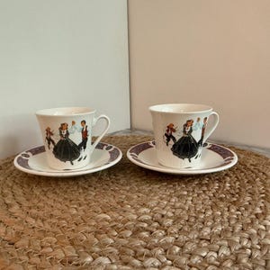 May include: Two white teacups and saucers with a decorative design of people dancing. The cups and saucers have a purple border. The cups have a handle and are sitting on a woven mat.