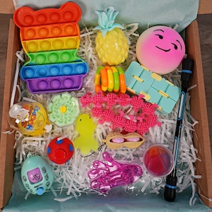 May include: A selection of fidget toys in a cardboard box. Includes a rainbow bear-shaped pop-it, a yellow pineapple, a pink emoji stress ball, and other sensory toys in various shapes and colours.
