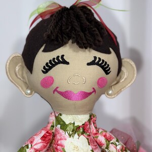 May include: A handmade doll with brown yarn hair, pink cheeks, and a pink smile. The doll is wearing a floral dress with pink and white roses.