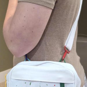以下が含まれることがあります： 白いクロスボディバッグ。控えめなエンボス加工のグリッドパターン、シルバーのジッパー開閉部、白いジャンプマンロゴのパッチが特徴です。バッグは赤、緑、青、オレンジのカラフルなペイントスプラッターで装飾されており、白いストラップが付いています。