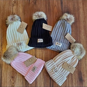 May include: Five ribbed knit beanies with faux fur pom-poms. Colors include cream, black, gray, pink, and tan. Each beanie has a folded brim and a rectangular tag with the brand name "UGG".
