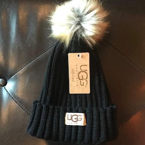 Könnte beinhalten: Eine schwarze Strickmütze mit einem Kunstfell-Bommel. Die Mütze hat ein geripptes Design und einen kleinen UGG-Logoaufnäher. Ein Geschenkkartenanhänger mit dem UGG Australia-Logo ist angebracht.