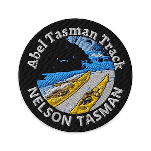 May include: A black and white embroidered patch with the text "Abel Tasman Track Nelson Tasman" in a circle. The patch features a blue and white image of a mountain range with two yellow kayaks in the foreground.