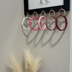 Puede incluir: Seis llaveros de silicona coloridos con anillos dorados colgados de ganchos. Los llaveros son en tonos de rosa, blanco y rojo. Los ganchos están unidos a un marco negro con un letrero blanco que dice "khan EST. 2020".