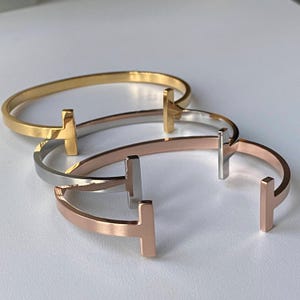 May include: Three open-ended bangles in gold, silver, and rose gold. Each bangle features a prominent T-shaped design on each end. The bracelets are displayed on a white surface, showcasing their polished finish and minimalist style.