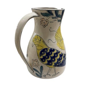 May include: A ceramic pitcher with a cream-colored base, featuring a hand-painted design of a yellow and blue bird. The pitcher has a curved handle and is adorned with floral and leaf motifs in blue and yellow. A decorative kitchenware item.