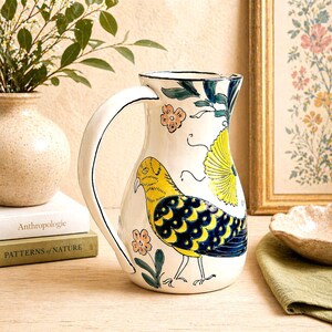 May include: A white ceramic pitcher with a large curved handle, decorated with a yellow and blue bird design, flowers, and green leaves. The pitcher is on a wooden surface, with books and a vase in the background.