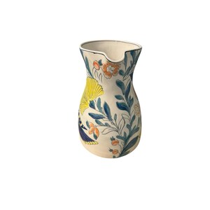 May include: A ceramic pitcher with a cream-colored base, adorned with a floral pattern in yellow, orange, and blue. The pitcher has a spout and a wide opening, suitable for serving beverages. The design includes stylized flowers and leaves.