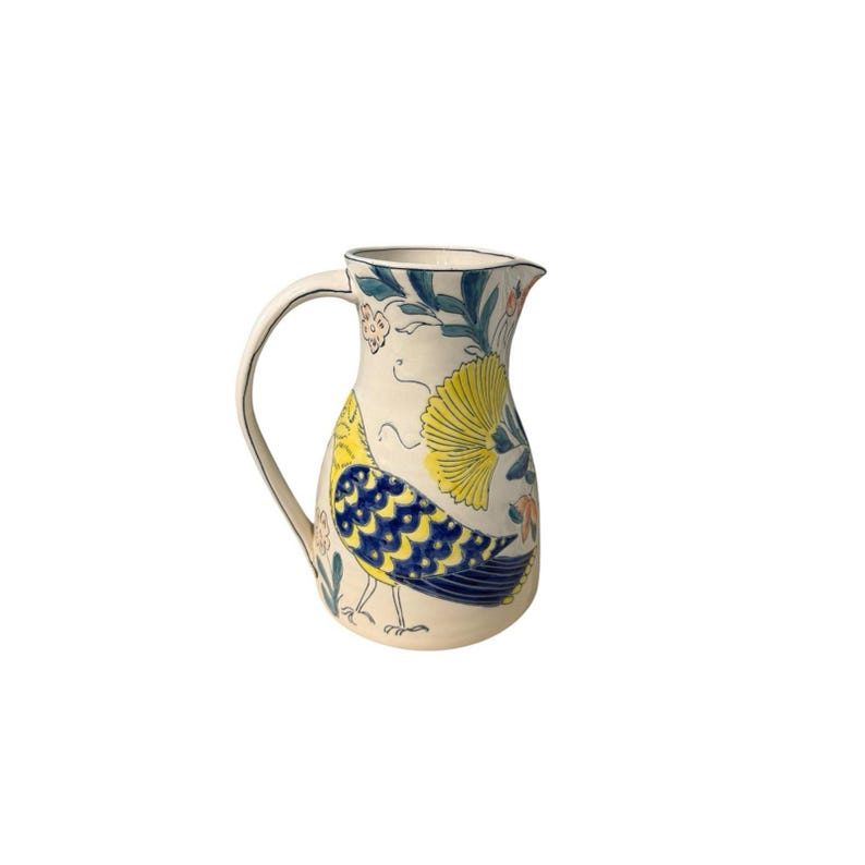 May include: Cream-colored ceramic pitcher with a curved handle and a spout. The pitcher is decorated with a yellow and blue bird design, along with floral and leaf patterns in blue, orange, and green. The pitcher is a decorative item.