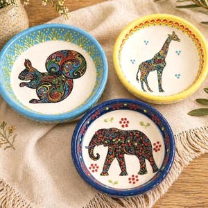 Könnte beinhalten: Drei dekorative Keramikschalen mit Tiermotiven. Eine Schale zeigt ein Eichhörnchen, eine andere eine Giraffe und die dritte einen Elefanten. Jede Schale hat ein farbenfrohes Muster und einen kontrastierenden Rand.