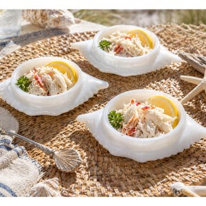 May include: Three white crab-shaped serving dishes filled with crab salad, lemon slices, and parsley. The dishes are on a woven mat, with a seashell spoon and starfish nearby. The scene evokes a coastal theme.
