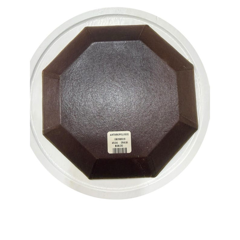 May include: A brown, octagonal dish inside a clear plastic container. The dish has a textured surface and a label with the text "ANTHROPOLOGIE". The dish is likely used for serving or display.