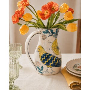 May include: A white ceramic pitcher with a large handle, decorated with a yellow and blue bird design and floral accents. The pitcher holds a bouquet of orange and yellow tulips. Two decorative glasses are in the background.